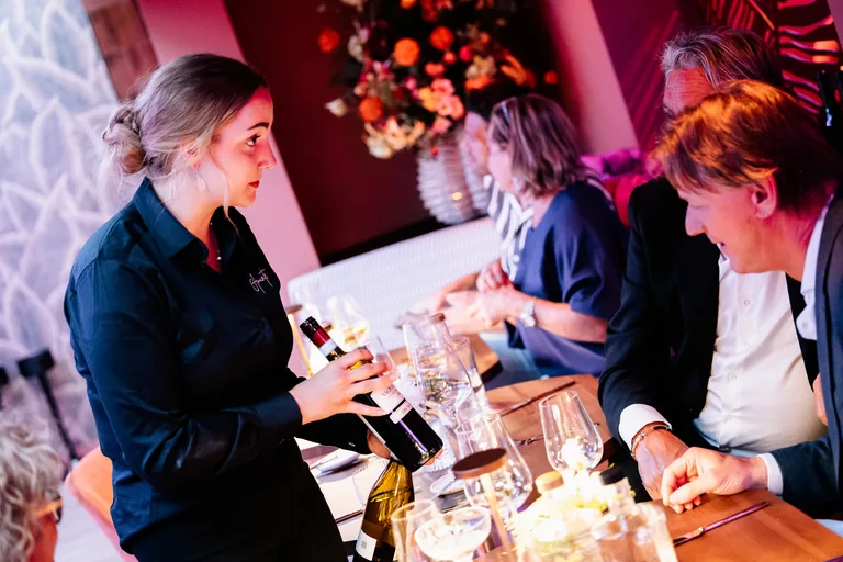 Een medewerker met een fles staat aan tafel bij gasten, met wijnglazen en warme verlichting.