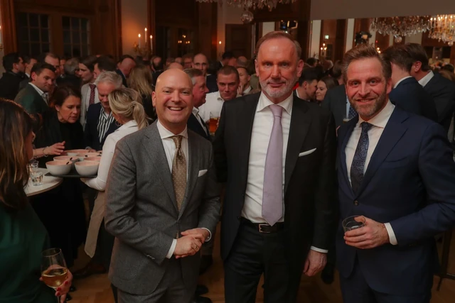 Drie gasten poseren samen met een levendige receptiezaal op de achtergrond.