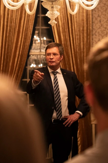 Een gast staat onder een kroonluchter en gebaart naar gasten op de voorgrond.