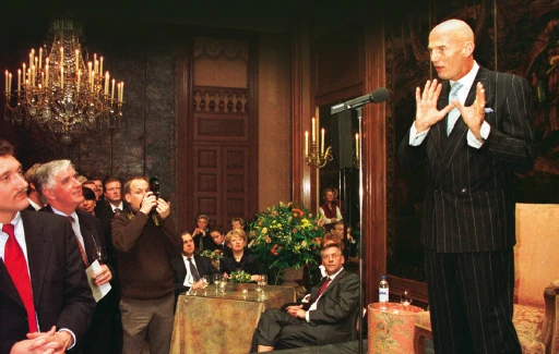 Pim Fortuyn spreekt bij een microfoon, met gasten op de achtergrond.