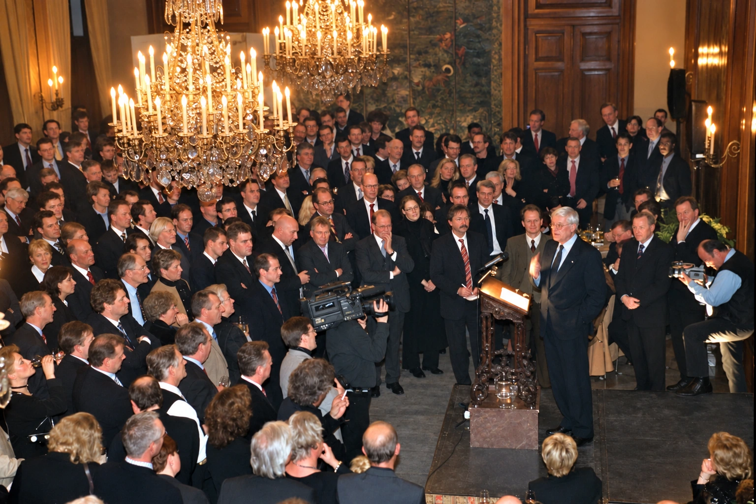 Historisch overzicht van een nieuwjaarsreceptie in de zaal van Paleis Het Loo, met gasten, kroonluchters en een spreker.