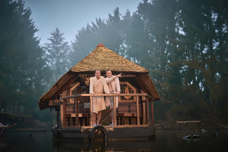 Twee gasten in badjassen staan voor een houten paviljoen met rieten dak aan het water.