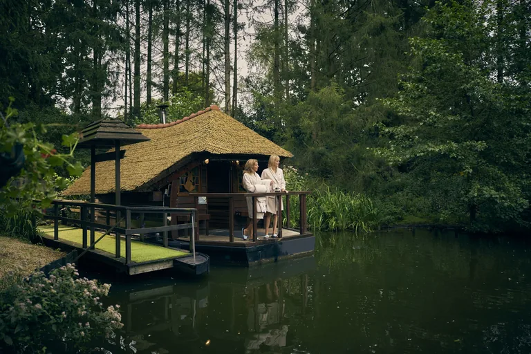 Een houten saunagebouw aan het water, omringd door bomen, met gasten op het terras.